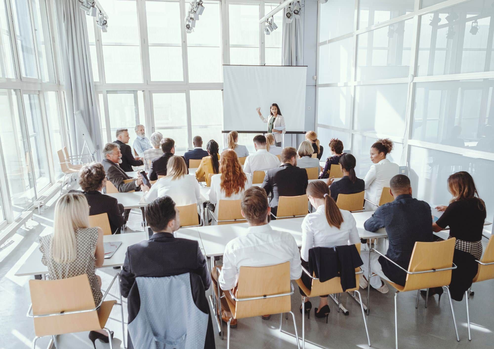 Teilnehmer eines Verkaufstrainings in Bonn, die auf eine Trainerin hören, die Verkaufsstrategien erklärt. Das Bild zeigt einen modernen Seminarraum und eine Gruppe von Personen, die an einem Seminar für Verkaufstraining teilnehmen.