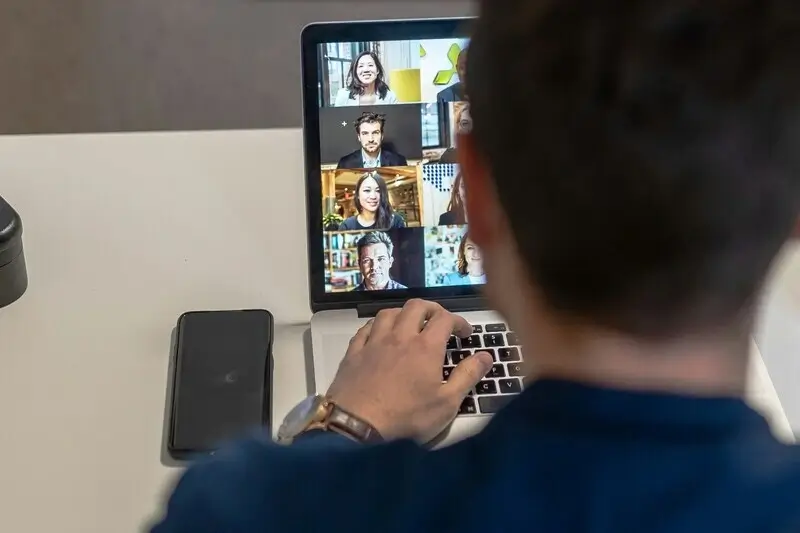 Hinterkopf eines Mitarbeiters, der an einem Online-Meeting teilnimmt, während auf seinem Laptop mehrere Videokonferenz-Teilnehmer zu sehen sind – ideal für ein E-Book über digitale Kommunikation.